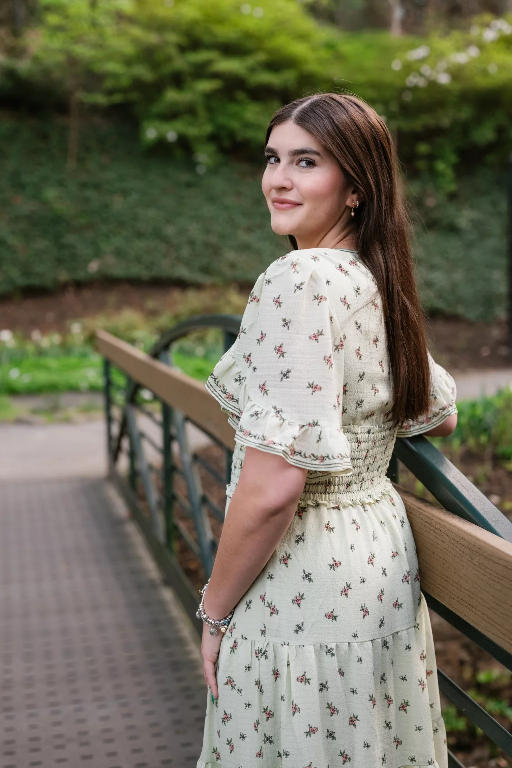Alivia's senior photos taken Downtown, Greenville, SC: A young woman with long brown hair, wearing a light-colored floral dress, stands on a bridge and looks back over her shoulder, smiling slightly. Lush greenery is visible in the background.