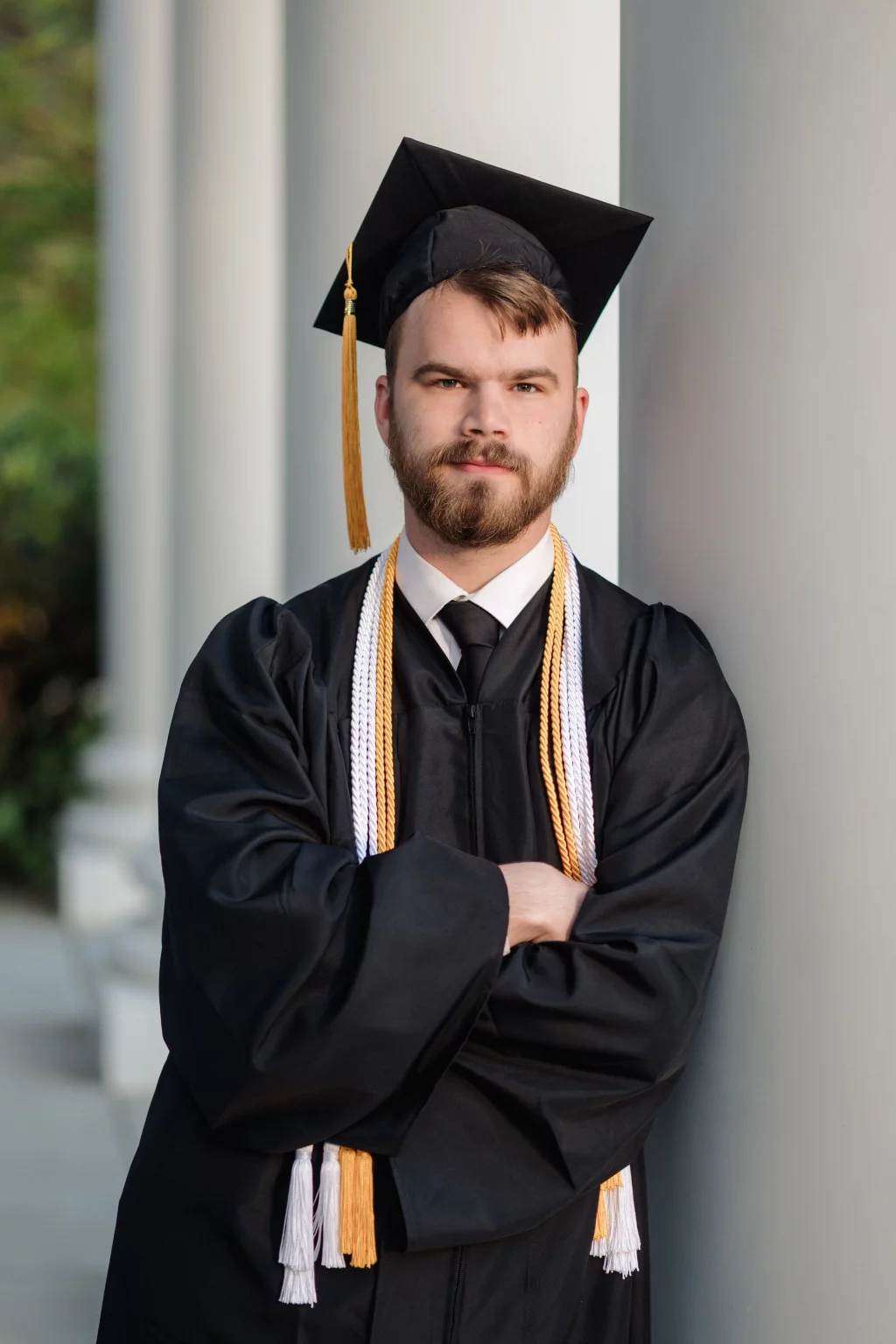Patrick Henry College & Liberty University graduation photos.: A young man in a black graduation cap and gown with honor cords stands with arms crossed, leaning against a column.