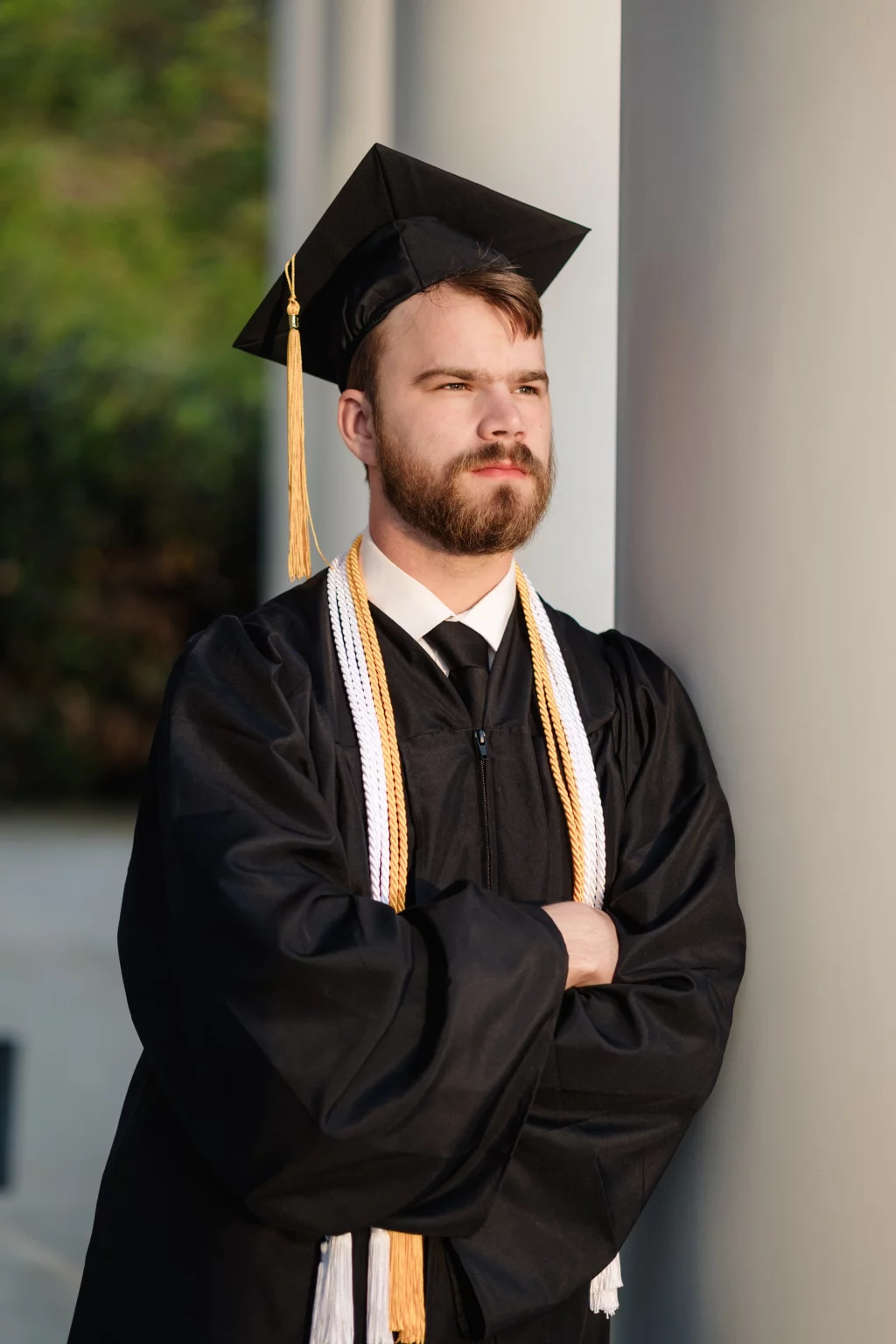 Patrick Henry College & Liberty University graduation photos.: Young man in a graduation cap and gown with honor cords, standing with arms crossed and leaning against a column, looking thoughtfully into the distance.