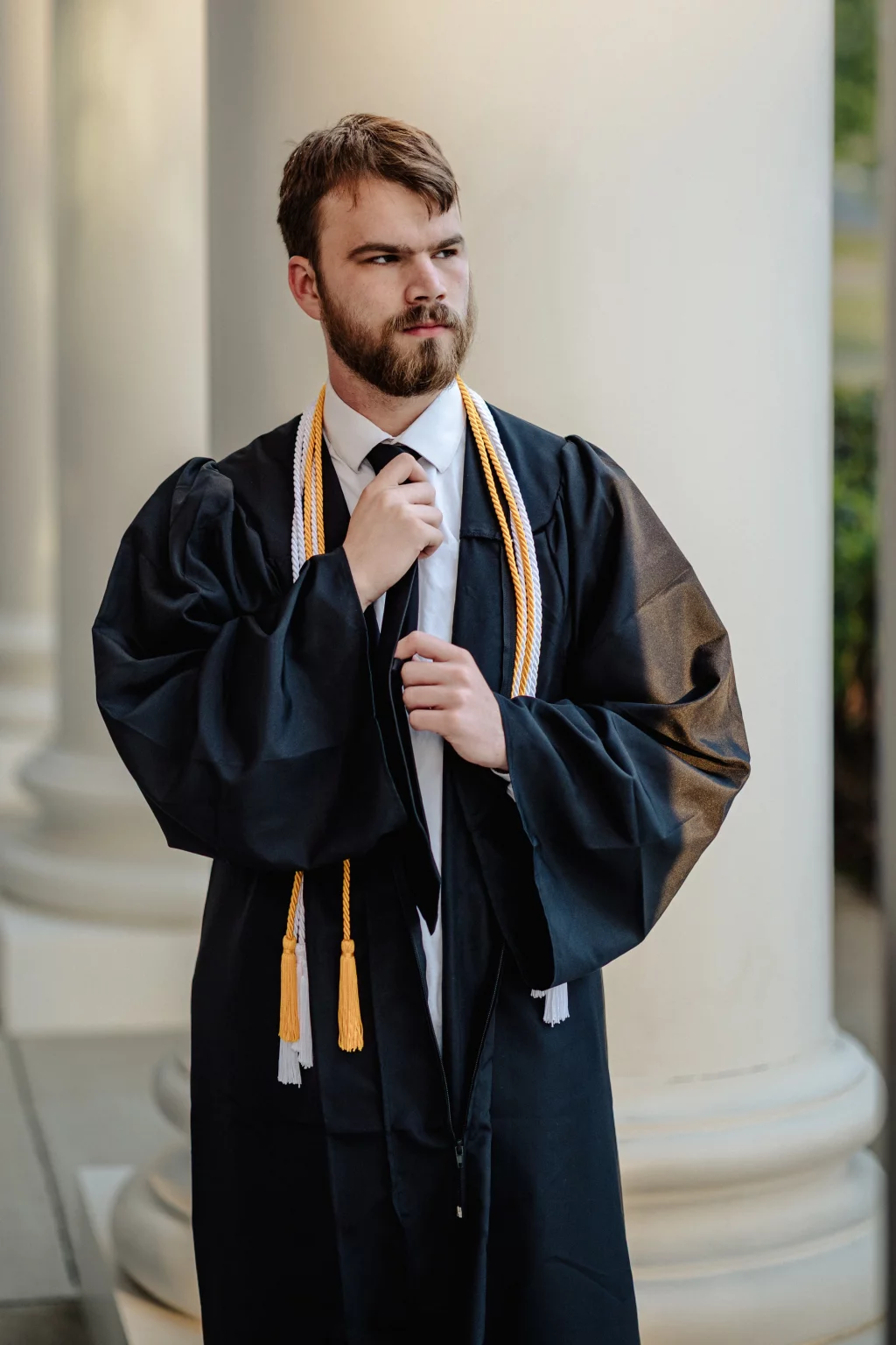 Patrick Henry College & Liberty University graduation photos.: A young man in a graduation gown and honor cords adjusts his tie while standing in front of large white columns.
