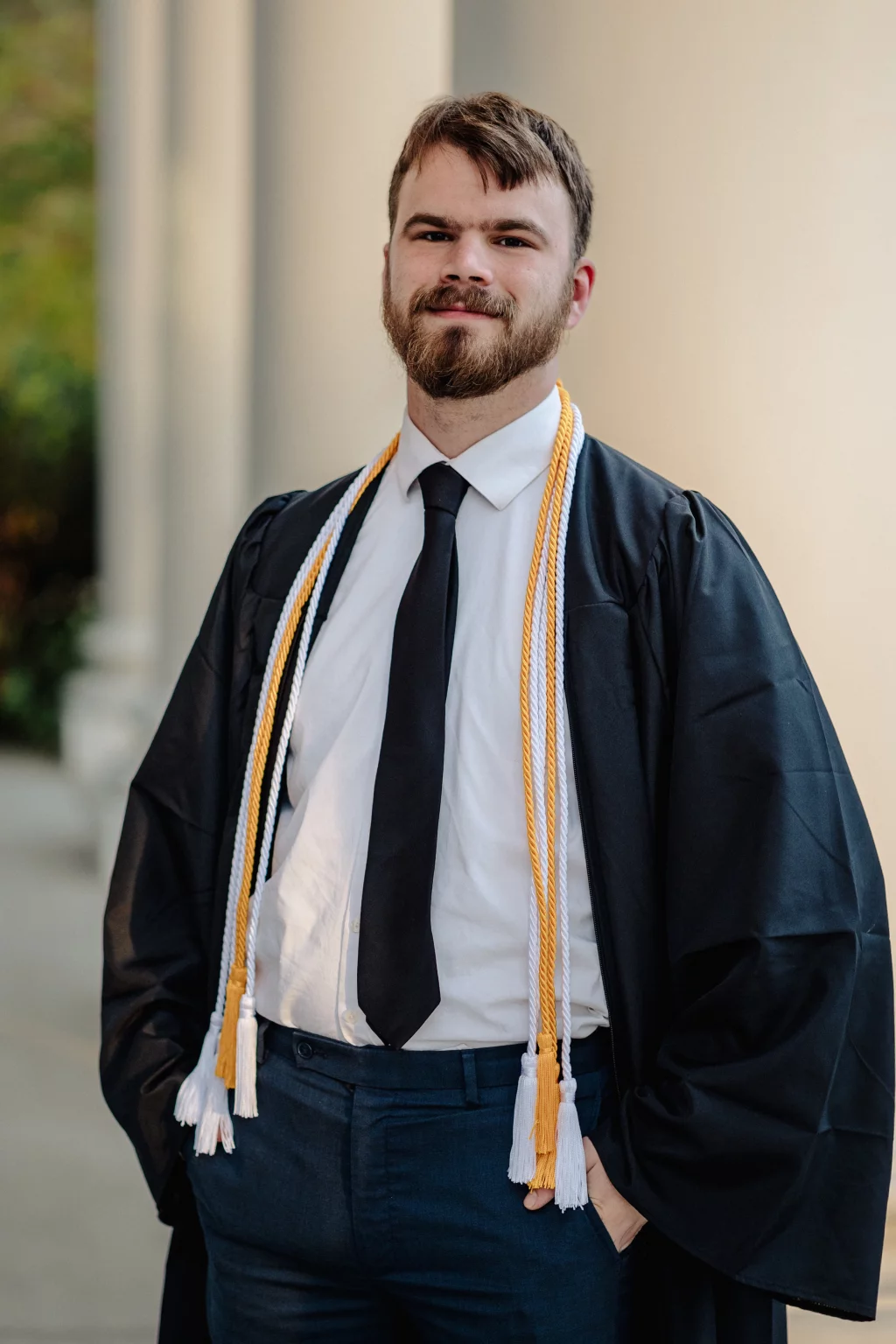 Patrick Henry College & Liberty University graduation photos.: A young man wearing a graduation gown, honor cords, white shirt, and black tie, standing outdoors and smiling confidently.