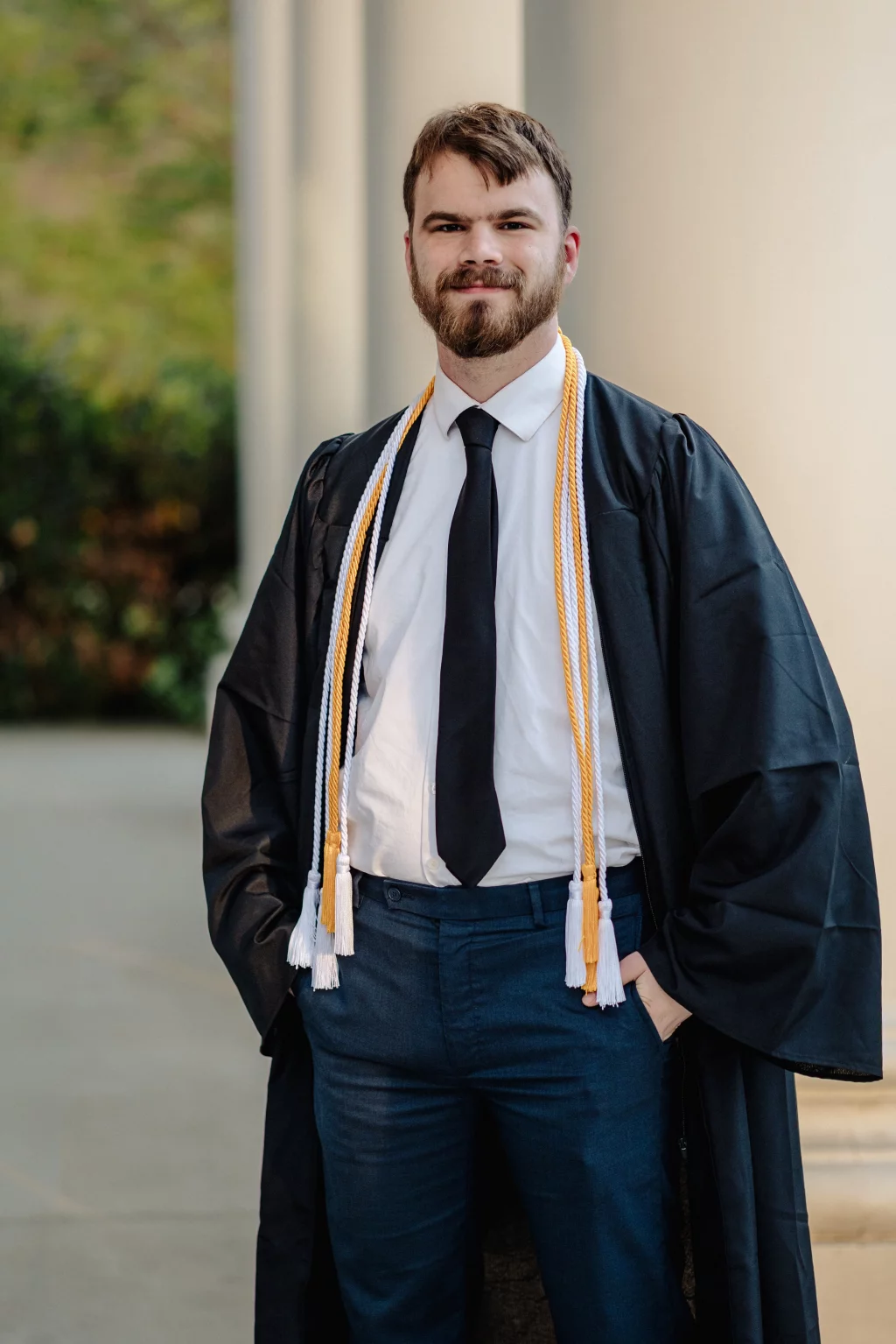 Patrick Henry College & Liberty University graduation photos.: A young man in a graduation gown with honor cords stands outdoors, smiling with his hands in his pockets.