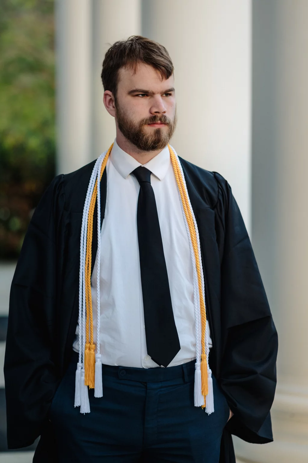 Patrick Henry College & Liberty University graduation photos.: A person wearing a graduation gown, honor cords, a white shirt, and a black tie stands in front of large columns, looking to the side.