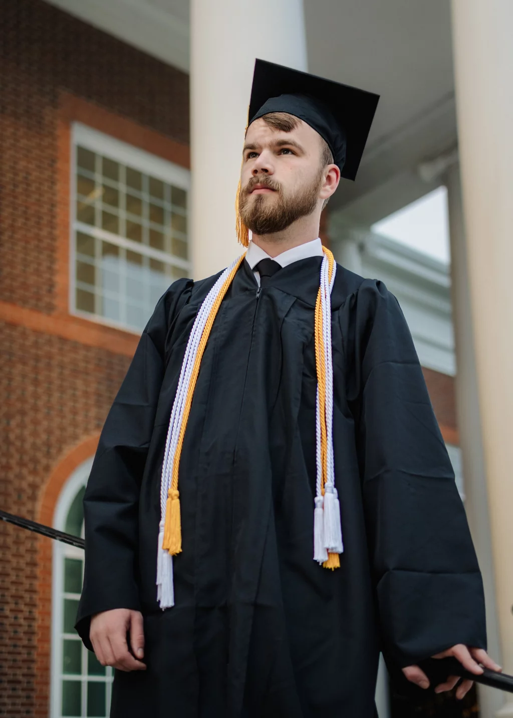 Patrick Henry College & Liberty University graduation photos.: A graduate in a black cap and gown with honor cords stands outside in front of a brick building and white columns, looking ahead confidently.