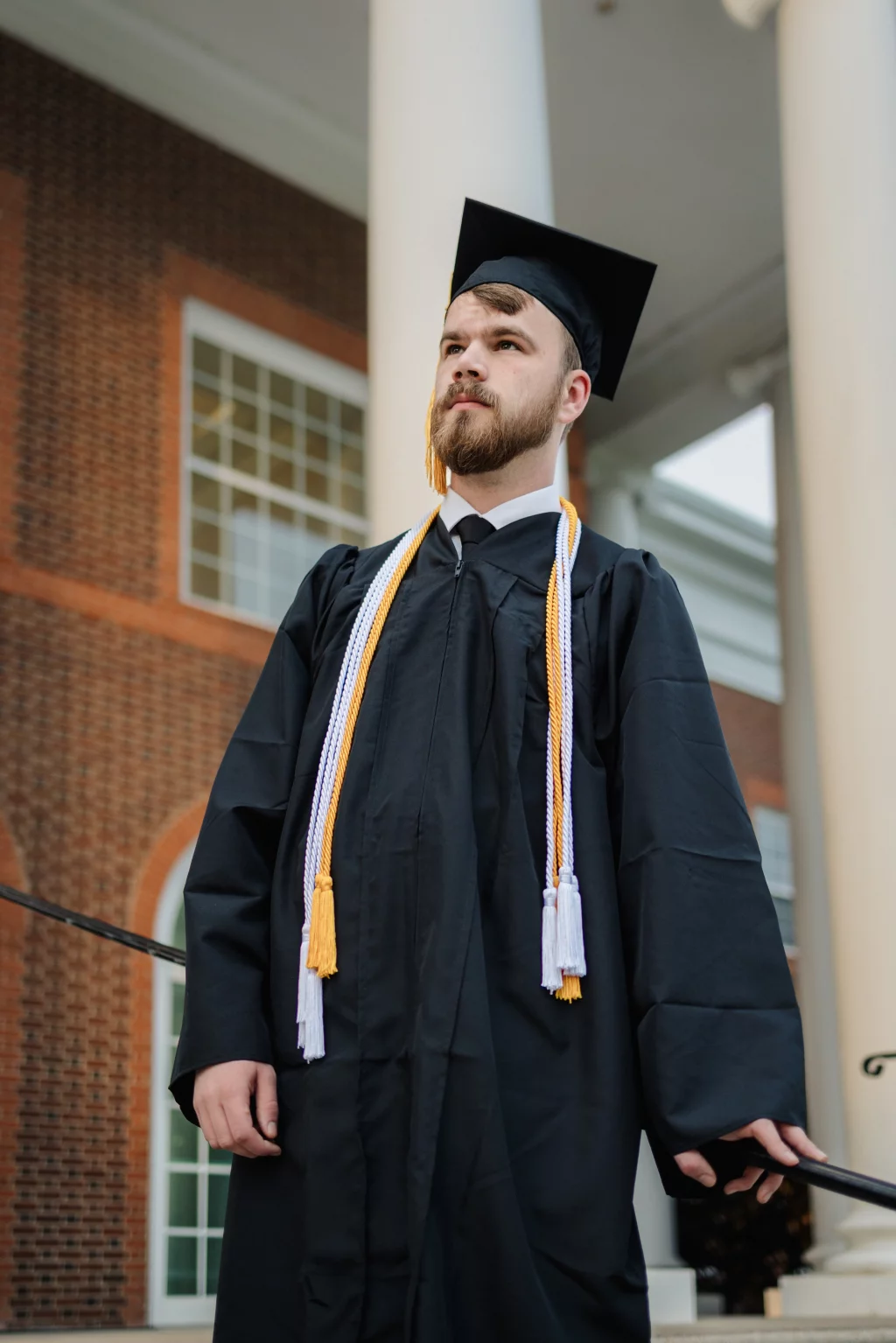 Patrick Henry College & Liberty University graduation photos.: A graduate wearing a black cap and gown with honor cords stands in front of a building with brick walls, large white columns, and tall windows.