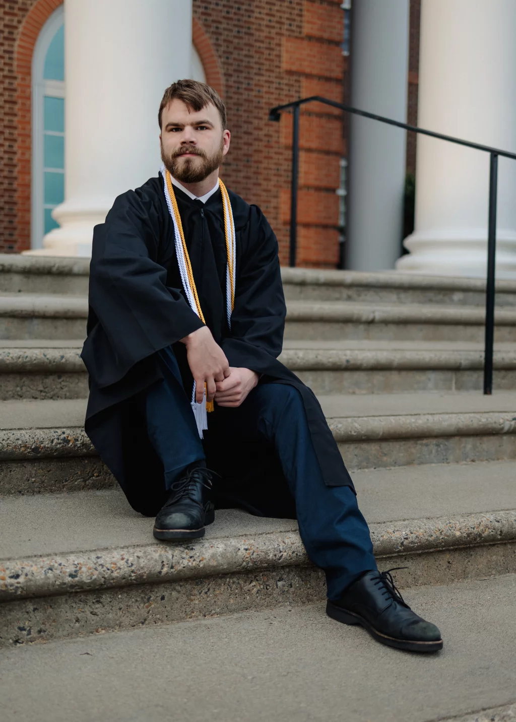 Patrick Henry College & Liberty University graduation photos.: A young man in a graduation gown and honor cords sits on outdoor steps in front of a building with large white columns and brick walls.