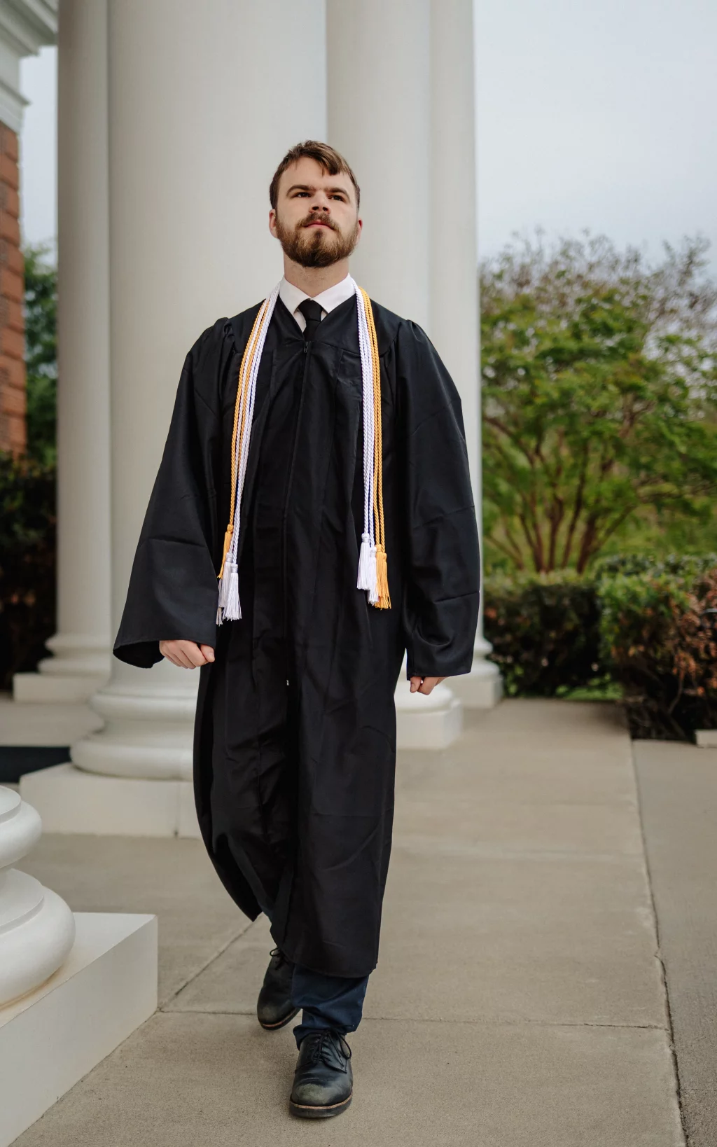Patrick Henry College & Liberty University graduation photos.: A young man in a black graduation gown and honor cords walks confidently on a sidewalk in front of large white columns, with greenery in the background.