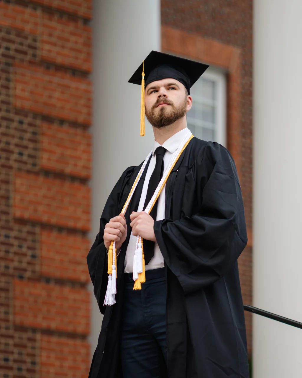 Patrick Henry College & Liberty University graduation photos.: A graduate wearing a black cap and gown with honor cords stands confidently in front of a brick building.