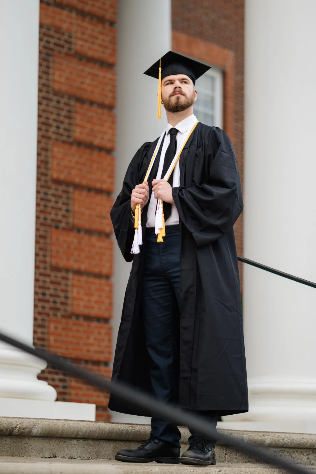 Patrick Henry College & Liberty University graduation photos.: A person in a graduation cap and gown stands on stone steps in front of a brick building with white columns, looking upward confidently.