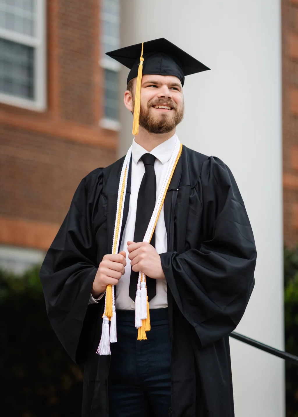 Patrick Henry College & Liberty University graduation photos.: A young man in a graduation cap and gown, wearing honor cords, stands outside in front of a brick building, smiling and looking away from the camera.