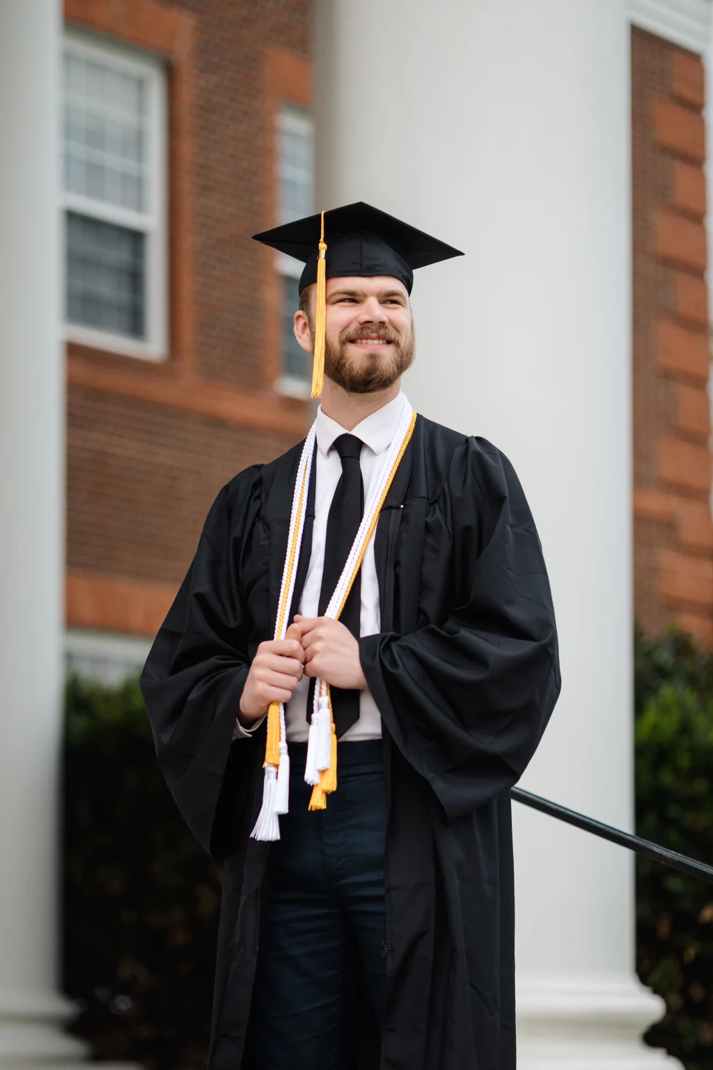Patrick Henry College & Liberty University graduation photos.: A young man in a graduation cap and gown stands smiling in front of a brick building with white columns, holding honor cords.