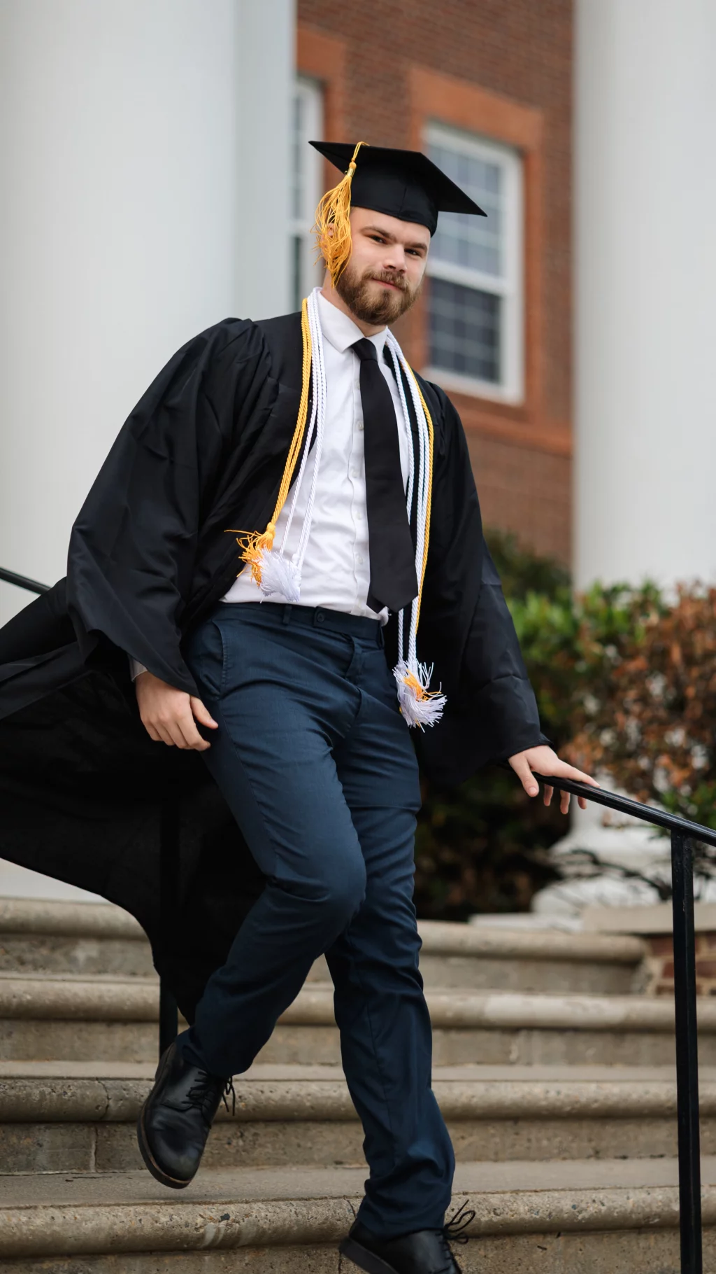 Patrick Henry College & Liberty University graduation photos.: A young man wearing a graduation cap, gown, and honor cords walks down outdoor steps, holding a handrail, with a brick building and columns in the background.