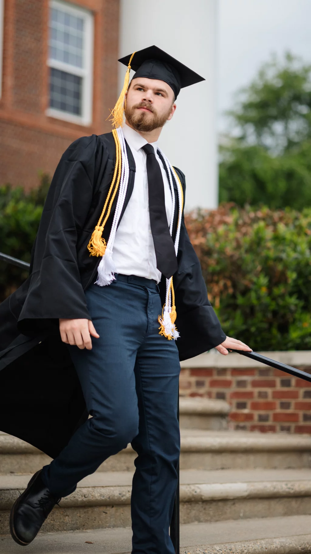 Patrick Henry College & Liberty University graduation photos.: A young man in a graduation cap and gown with honor cords walks down outdoor steps, holding a handrail. Brick building and greenery are visible in the background.