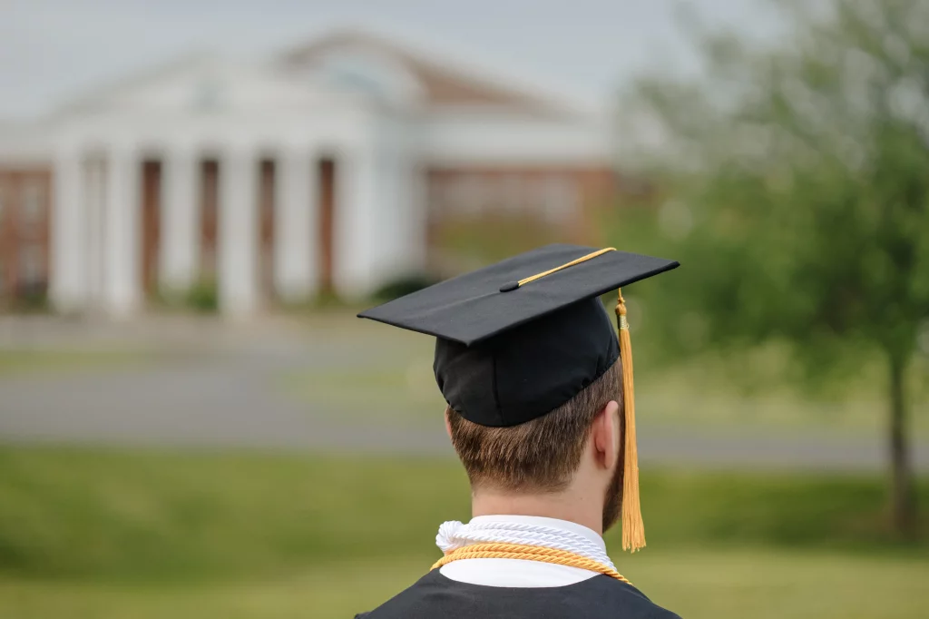 Patrick Henry College & Liberty University graduation photos.: A graduate wearing a cap and gown is seen from behind, standing outdoors with a blurred building featuring white columns in the background.