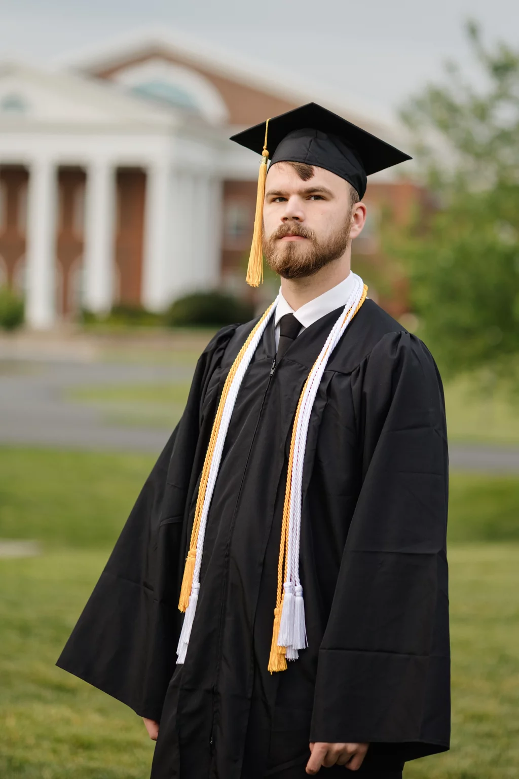 Patrick Henry College & Liberty University graduation photos.: A young man in a black graduation cap and gown with honor cords stands outside on grass, with a blurred building featuring white columns in the background.
