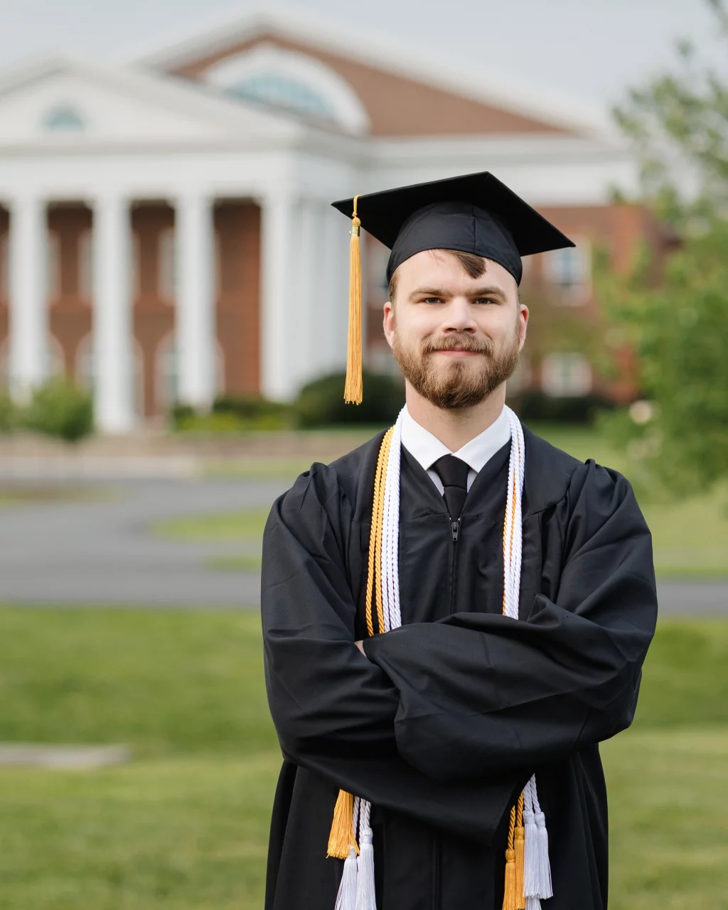 Patrick Henry College & Liberty University graduation photos.: Young man in a graduation cap and gown with honor cords stands outdoors with arms crossed, smiling, in front of a large brick building with white columns.