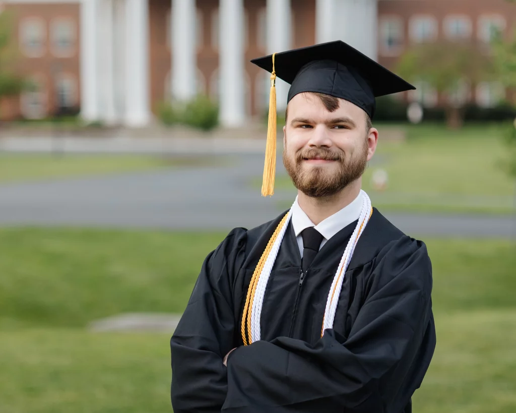 Patrick Henry College & Liberty University graduation photos.: A young man wearing a black graduation cap and gown with honor cords stands smiling outdoors, with a blurred building and green lawn in the background.