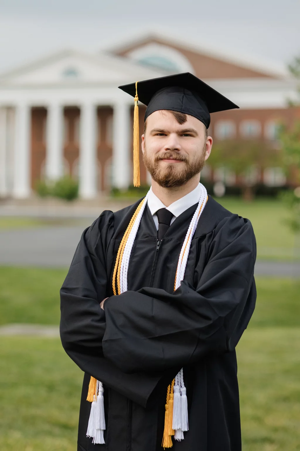 Patrick Henry College & Liberty University graduation photos.: A graduate wearing a black cap and gown with honor cords stands outdoors in front of a building with white columns and red brick.