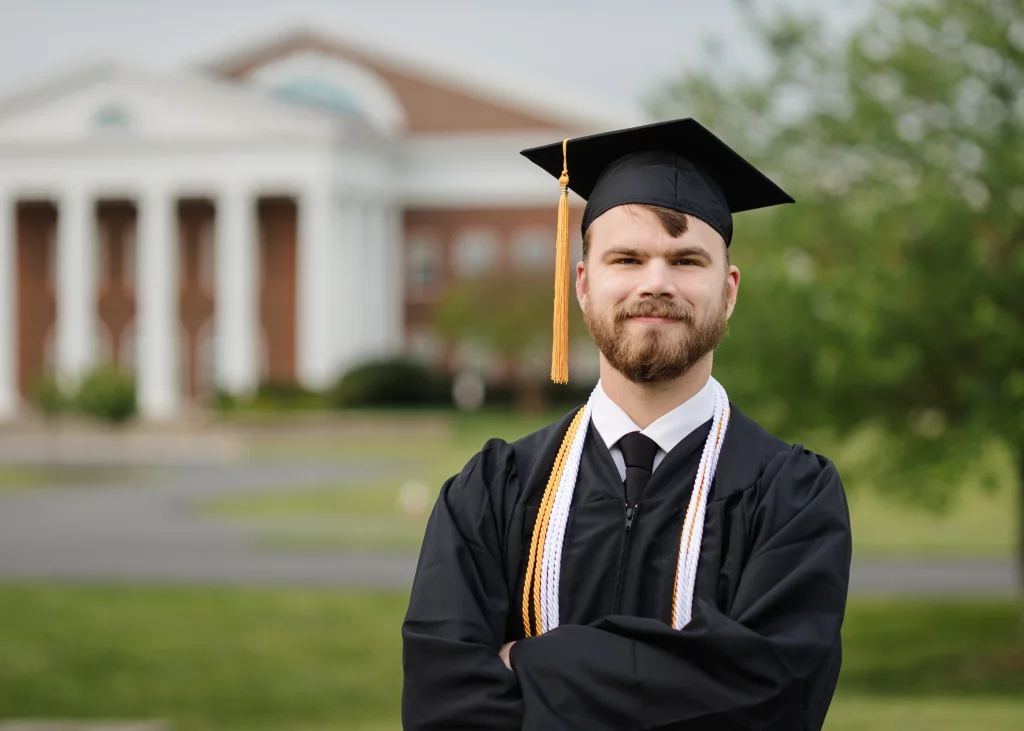 Patrick Henry College & Liberty University graduation photos.: A young man wearing a graduation cap and gown stands outdoors with arms crossed, smiling. A large building with white columns and green trees are in the background.