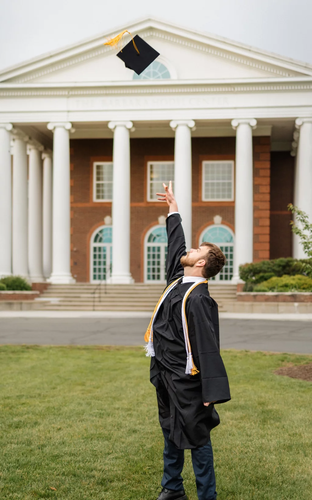 Patrick Henry College & Liberty University graduation photos.: A graduate in cap and gown tosses their mortarboard into the air in front of a building with white columns.