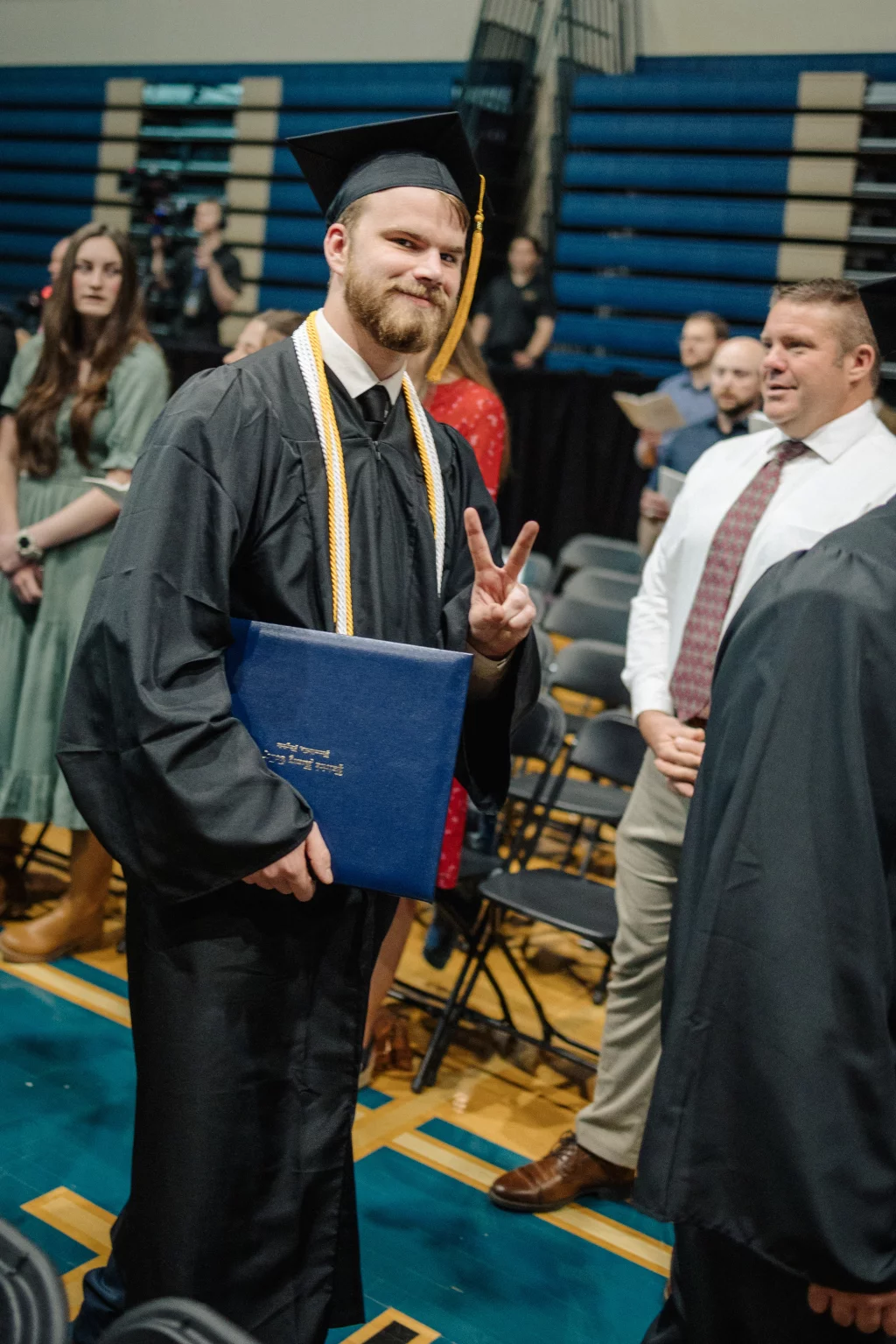 Patrick Henry College & Liberty University graduation photos.: A graduate in a black cap and gown holds a diploma folder and makes a peace sign, standing in a gymnasium with other people in the background.