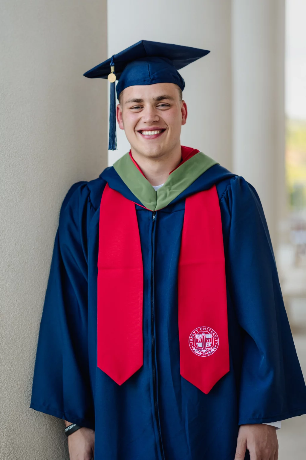 Patrick Henry College & Liberty University graduation photos.: A smiling graduate wearing a blue cap and gown with a green hood and red stole, standing against a light-colored wall. The stole features a university seal.