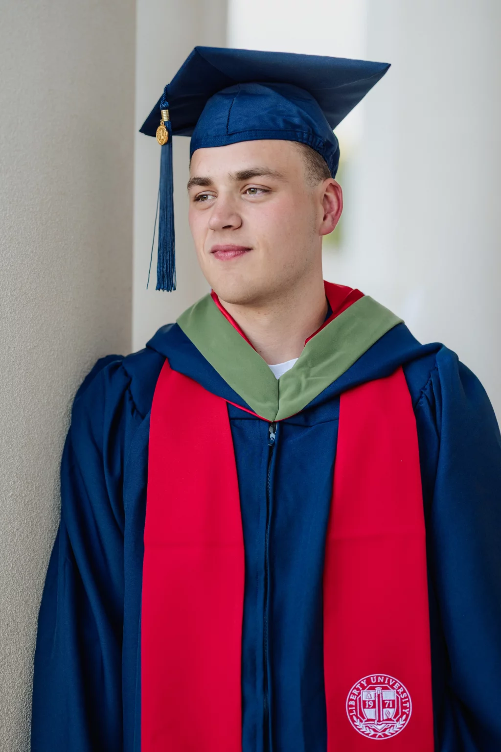 Patrick Henry College & Liberty University graduation photos.: A young man in a blue graduation cap and gown with a red stole and green hood stands against a wall. The stole features the Liberty University seal.