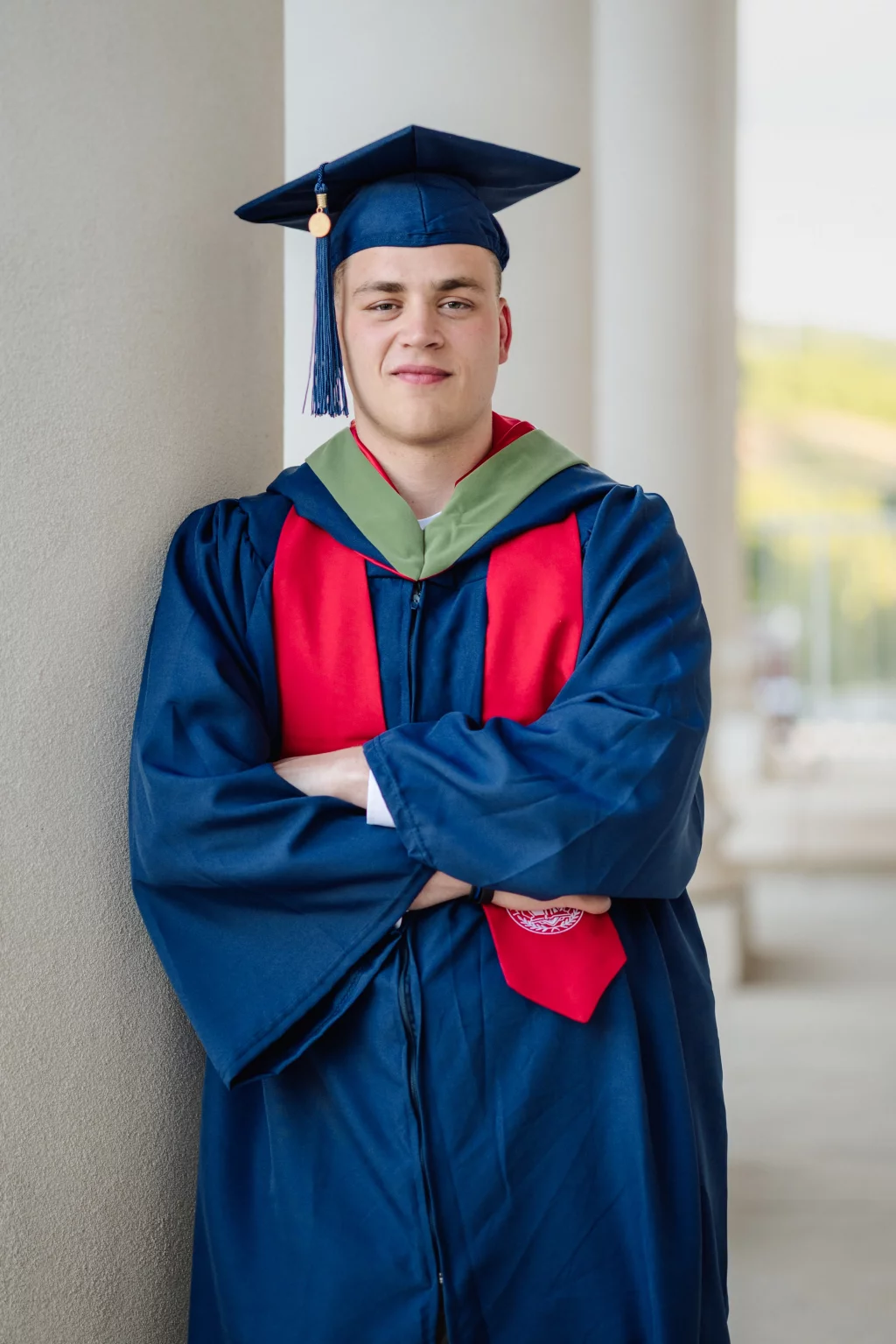Patrick Henry College & Liberty University graduation photos.: A young person in a blue graduation cap and gown with a red stole stands with arms crossed, leaning against a column.