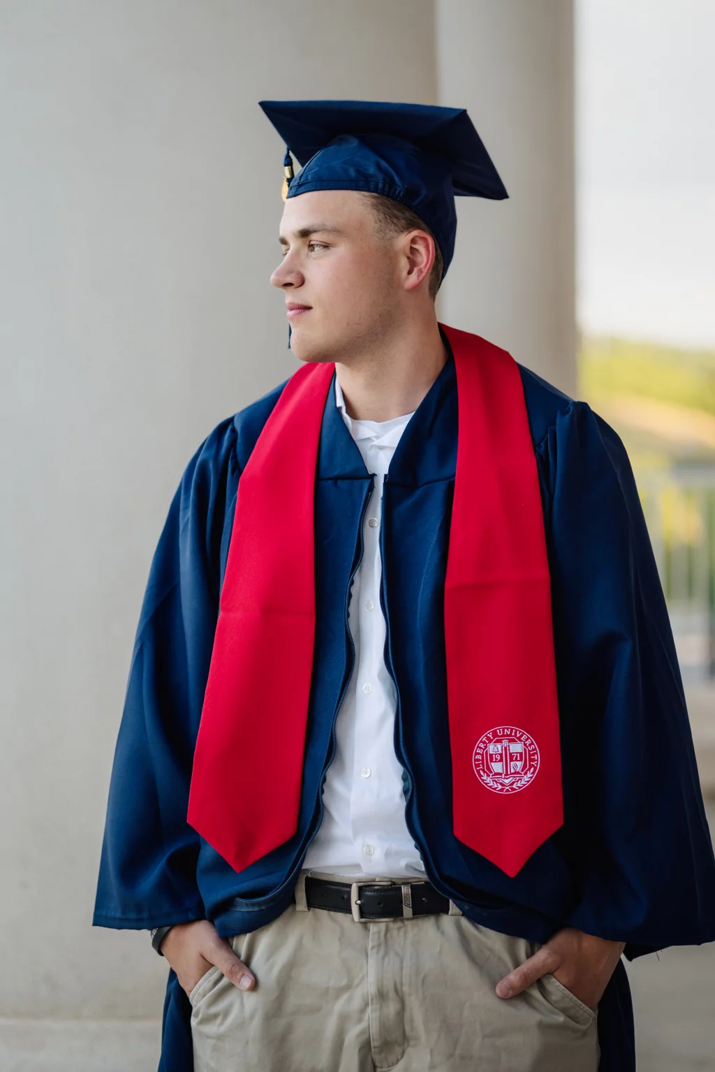 Patrick Henry College & Liberty University graduation photos.: A young man in a blue graduation gown and cap with a red stole stands with his hands in his pockets, looking to the side. The stole has a Liberty University emblem.