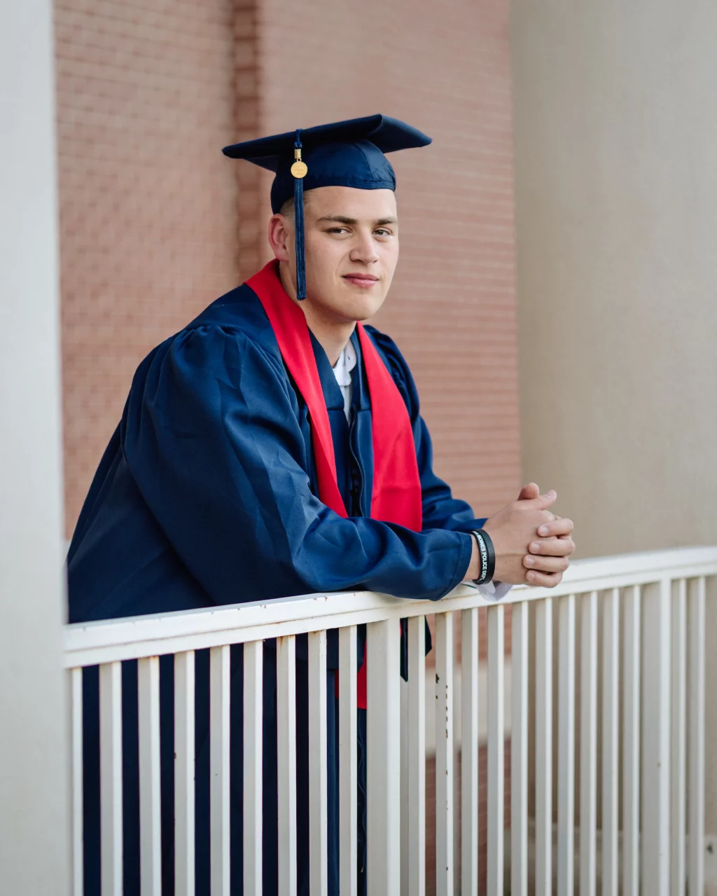 Patrick Henry College & Liberty University graduation photos.: A graduate in a navy blue cap and gown with a red stole leans on a white railing, posing for a photo.