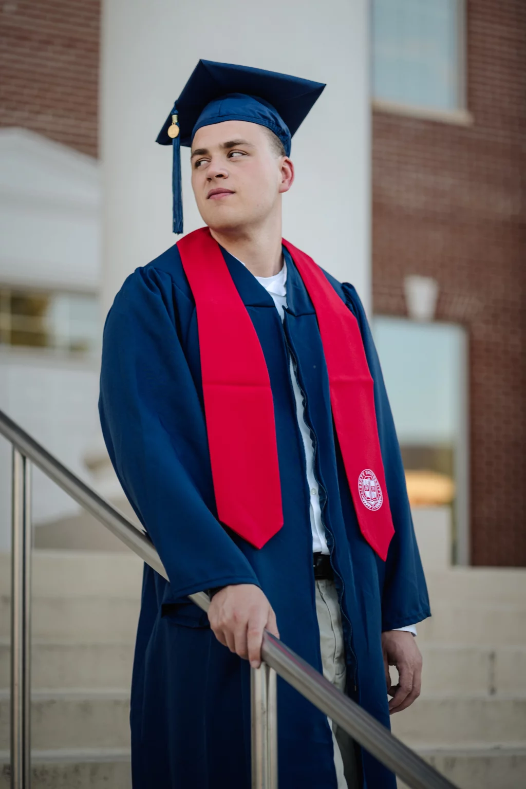 Patrick Henry College & Liberty University graduation photos.: A young man in a blue graduation gown and cap with a red stole stands on outdoor steps, holding a railing and looking to the side.