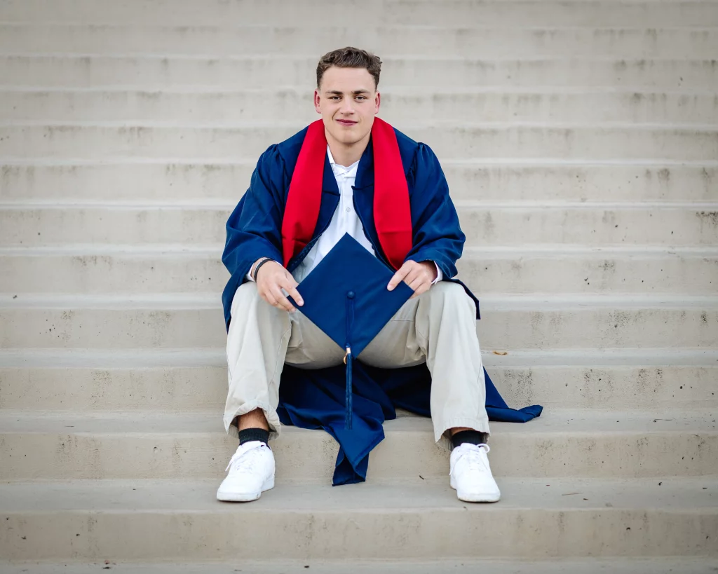 Patrick Henry College & Liberty University graduation photos.: A young man in a blue graduation gown with a red stole sits on outdoor concrete steps, holding a blue graduation cap and smiling at the camera.