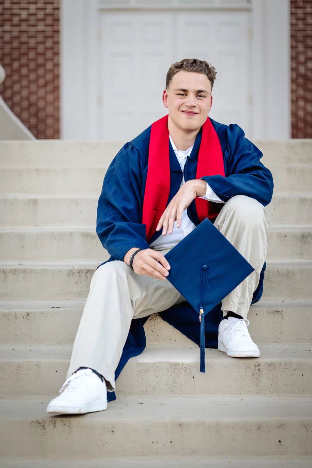 Patrick Henry College & Liberty University graduation photos.: Young man in a blue graduation gown with a red stole sits on outdoor steps, holding a blue graduation cap and smiling at the camera.