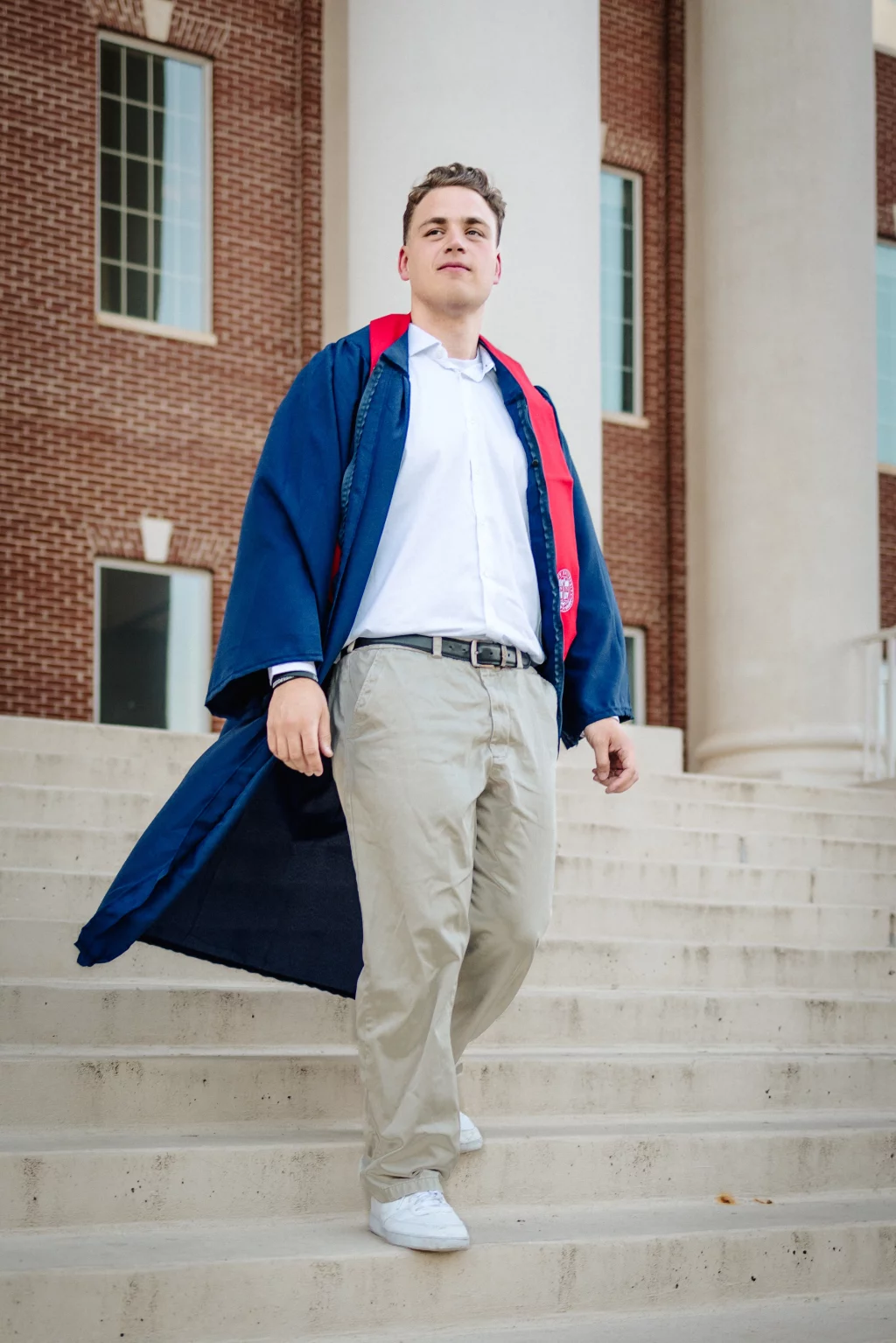 Patrick Henry College & Liberty University graduation photos.: A young man wearing a blue graduation gown with a red lining walks down concrete steps in front of a brick building with large white columns and windows.