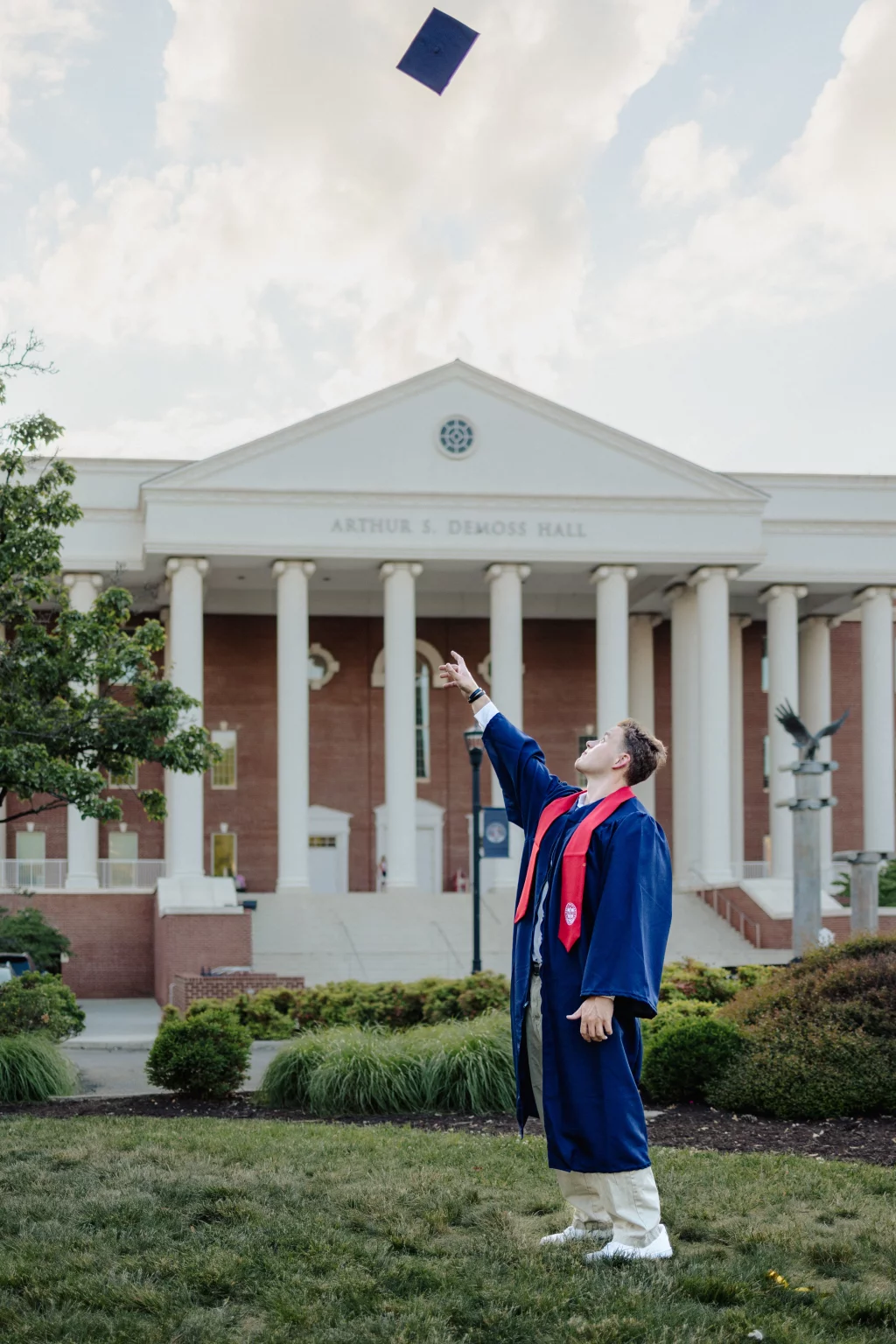 Patrick Henry College & Liberty University graduation photos.: A graduate in a blue gown and red stole stands on grass in front of Arthur S. Demoss Hall, pointing upward as a graduation cap is tossed into the sky.