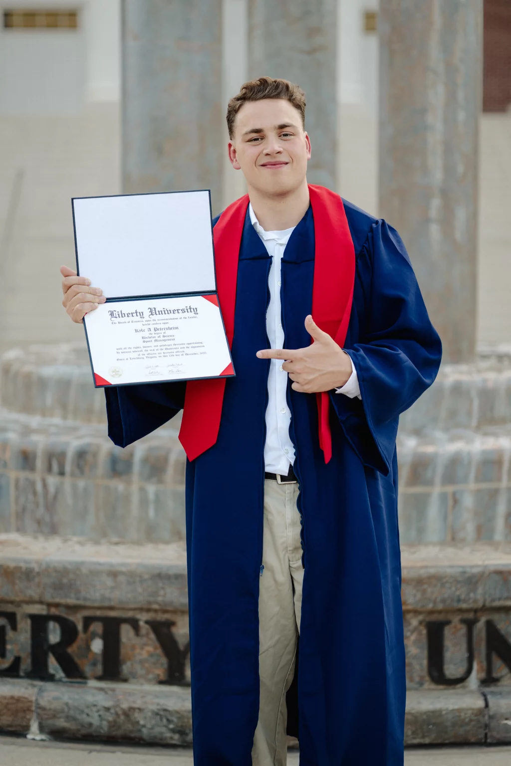 Patrick Henry College & Liberty University graduation photos.: A young man in a blue graduation gown with a red stole stands outdoors, holding up and pointing to his Liberty University diploma, smiling at the camera.