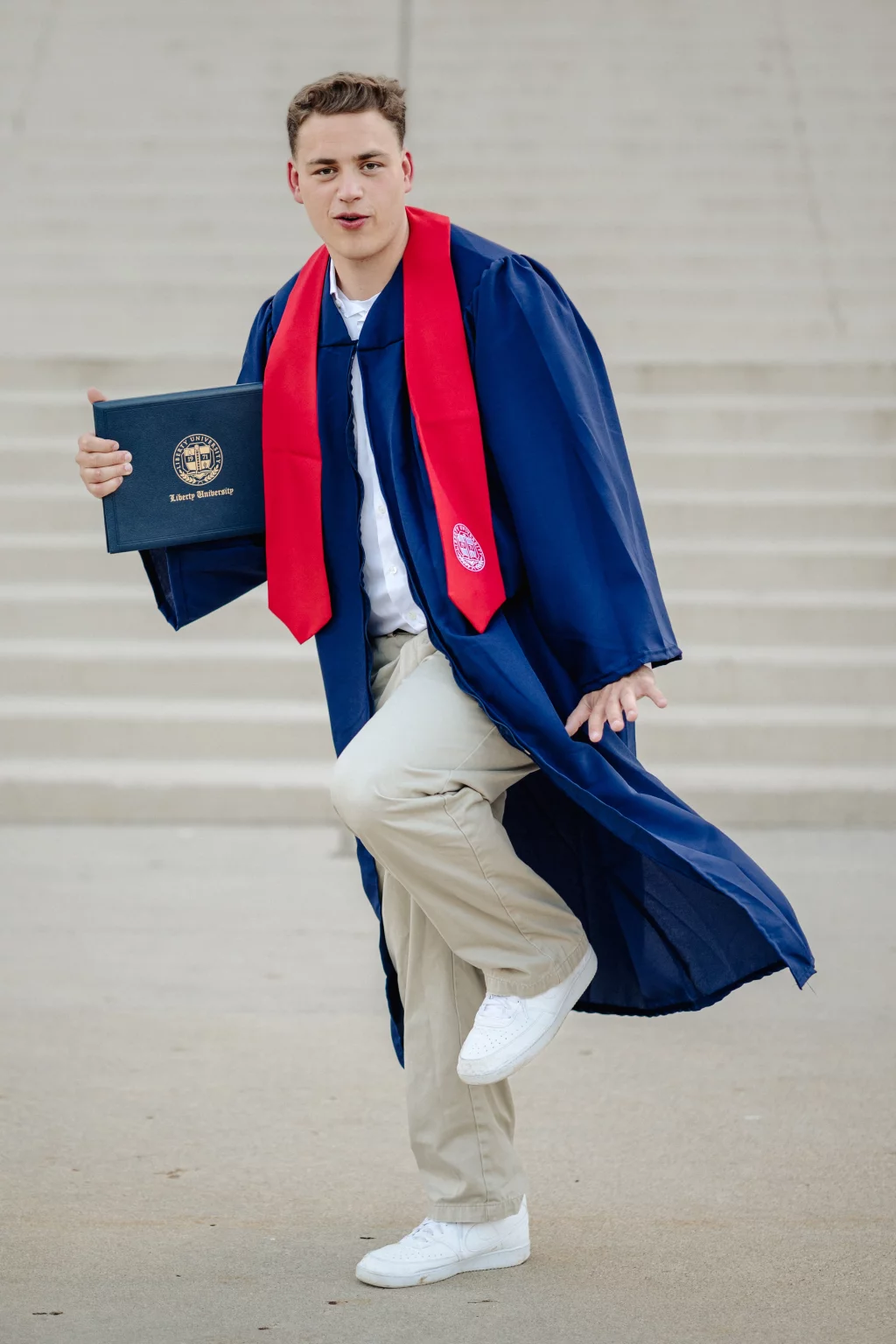 Patrick Henry College & Liberty University graduation photos.: A young man in a blue graduation gown and red stole poses playfully on one leg, holding a Liberty University diploma cover, with steps in the background.