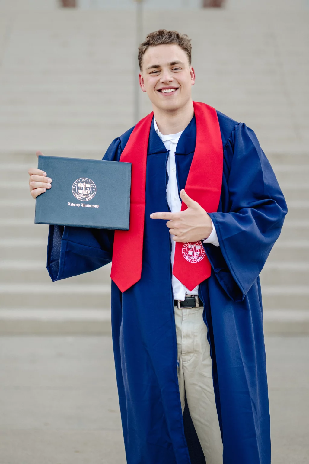 Patrick Henry College & Liberty University graduation photos.: Smiling graduate in a blue gown and red stole holding a Liberty University diploma cover and pointing at it, standing in front of steps.