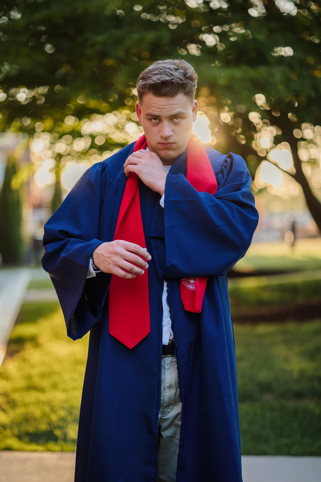 Patrick Henry College & Liberty University graduation photos.: A person wearing a blue graduation gown and red stole stands outdoors, adjusting the stole, with sunlight and trees in the background.