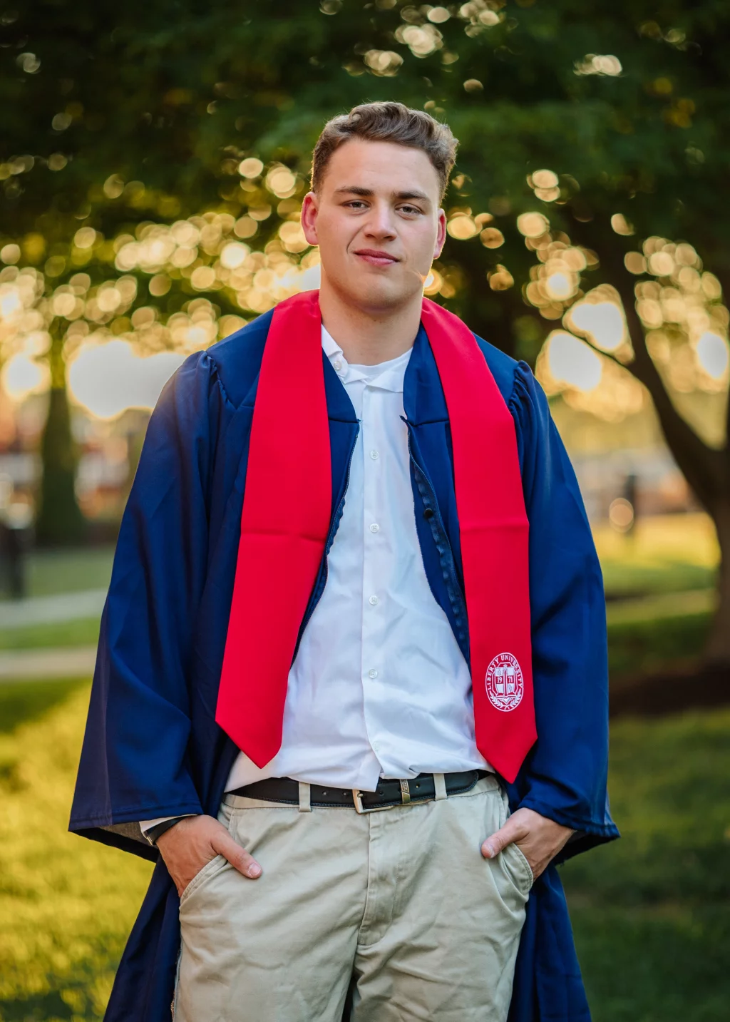 Patrick Henry College & Liberty University graduation photos.: Young man wearing a blue graduation gown, white shirt, and red stole, standing outdoors with hands in pockets; trees and sunlight are blurred in the background.