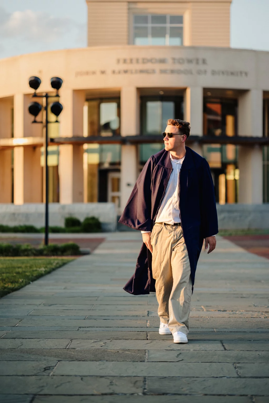 Patrick Henry College & Liberty University graduation photos.: A man wearing sunglasses and a dark graduation gown walks on a paved path in front of a building with "Freedom Tower" and "John W. Rawlings School of Divinity" written above the entrance.
