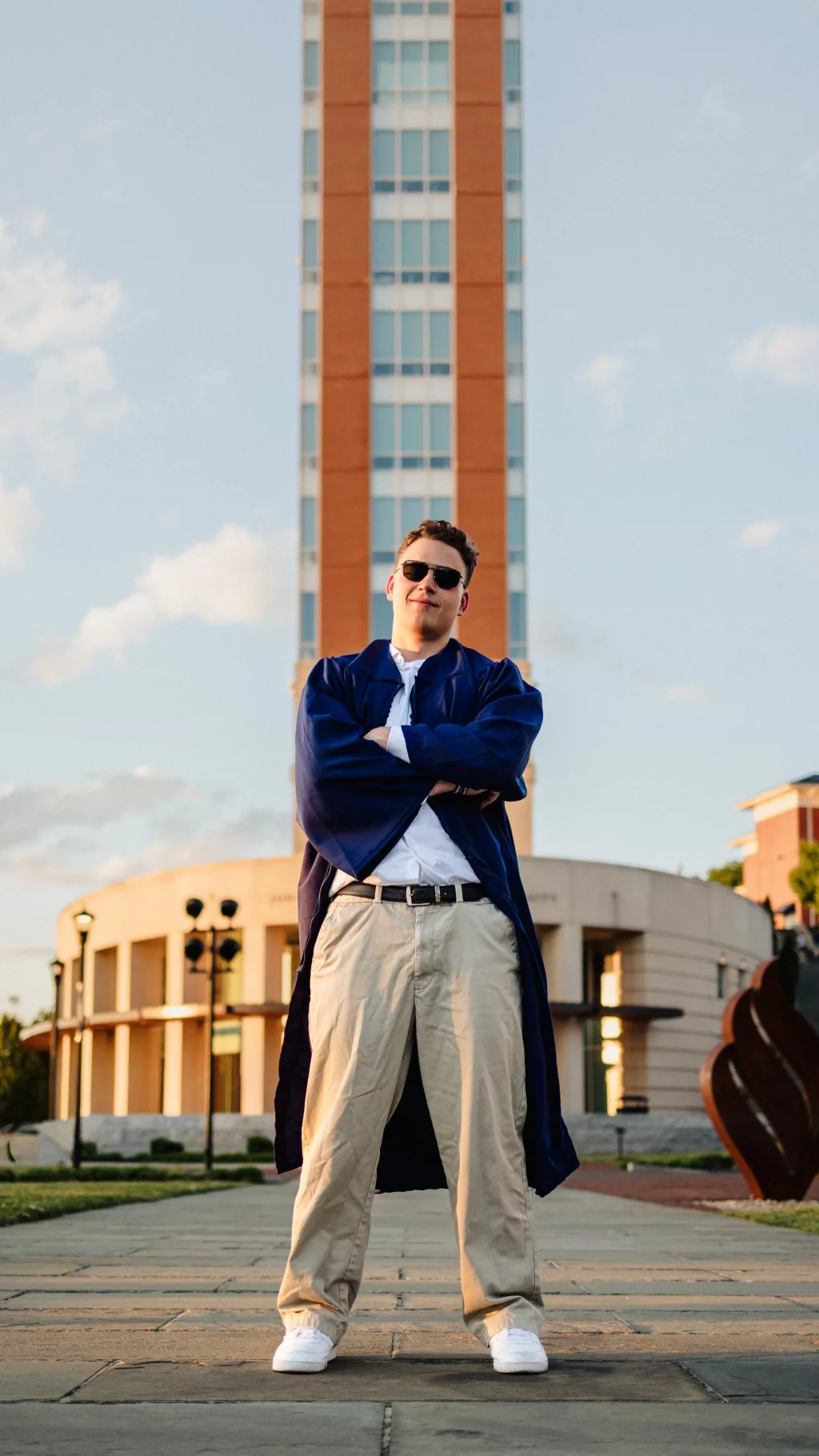 Patrick Henry College & Liberty University graduation photos.: A person wearing sunglasses and a blue graduation gown stands confidently with arms crossed in front of a tall brick and glass building.