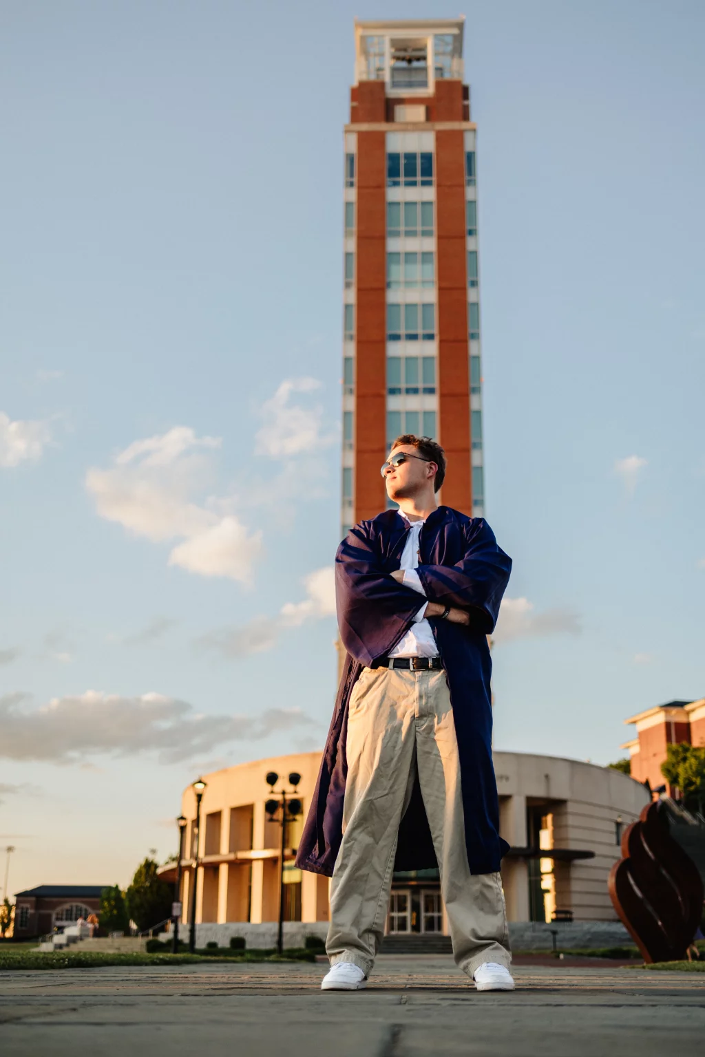 Patrick Henry College & Liberty University graduation photos.: A person in a blue graduation robe and sunglasses stands confidently with arms crossed in front of a tall brick clock tower, with a modern building and blue sky in the background.