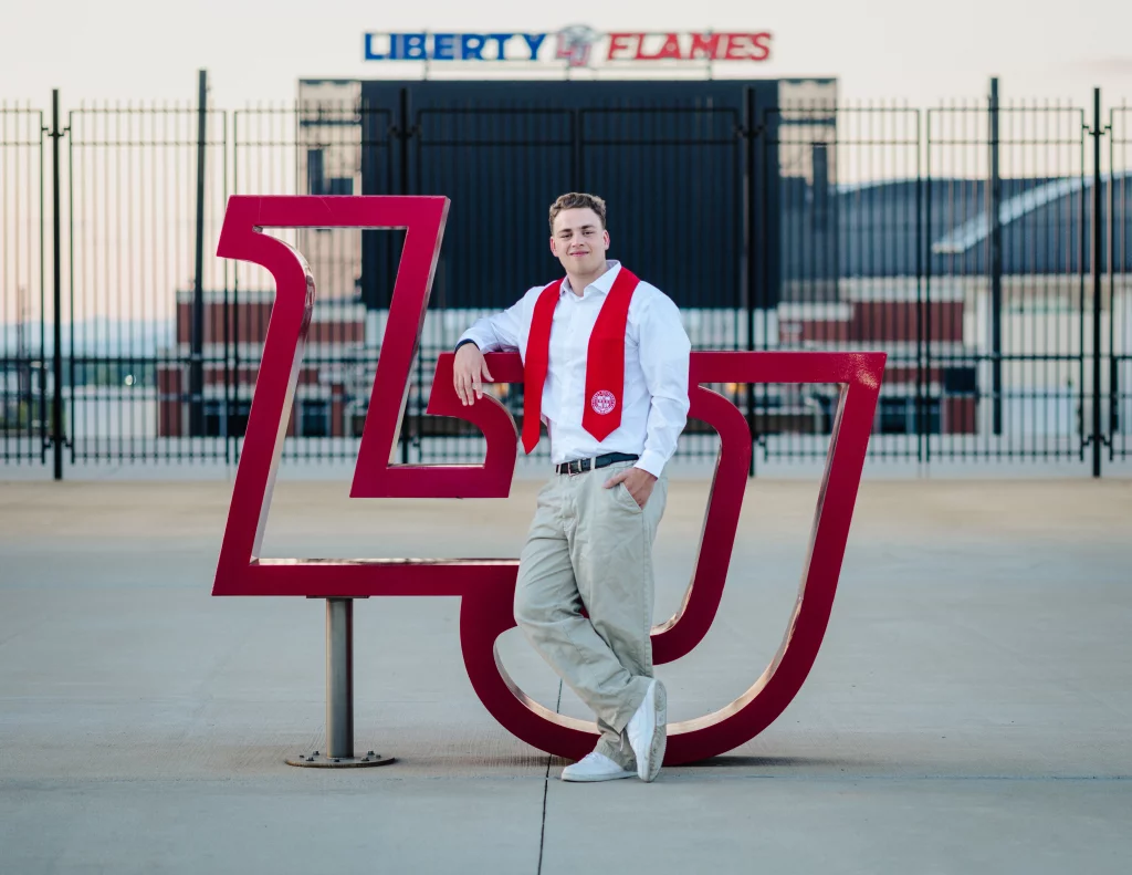 Patrick Henry College & Liberty University graduation photos.: A young man in a white shirt, khaki pants, and red graduation stole leans on a large red "LU" sculpture, with a stadium and a sign reading "Liberty Flames" in the background.