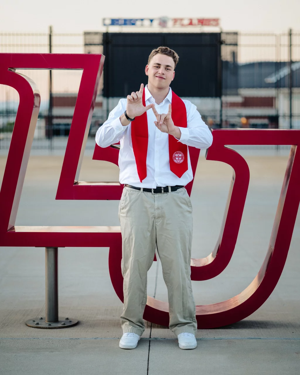 Patrick Henry College & Liberty University graduation photos.: Young man wearing a white shirt, khaki pants, and a red graduation stole stands in front of a large red "LU" sculpture, making a hand sign with both hands.