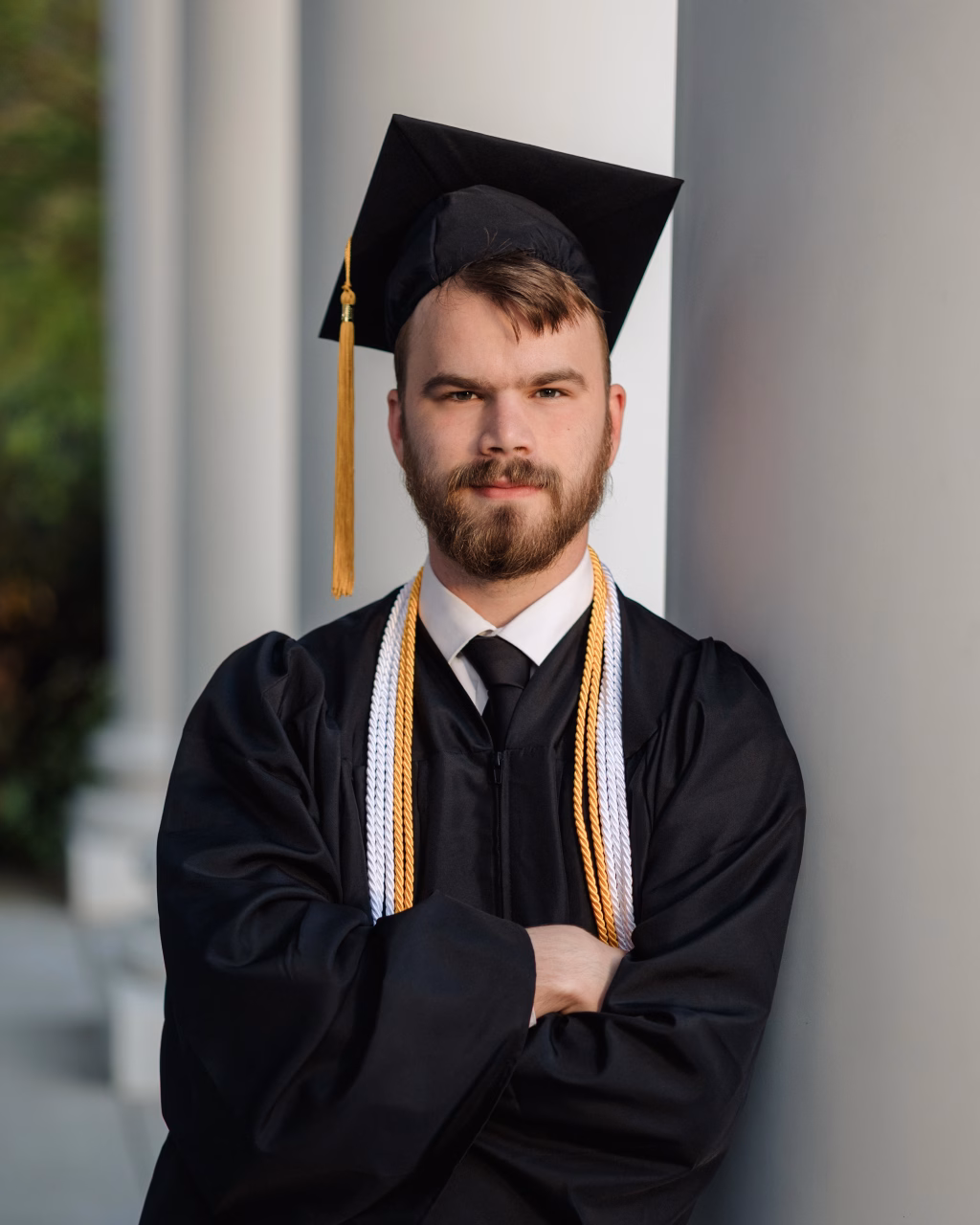 Patrick Henry College & Liberty University graduation photos.: A young man in a graduation cap and gown with honor cords stands with arms crossed, leaning against a column.