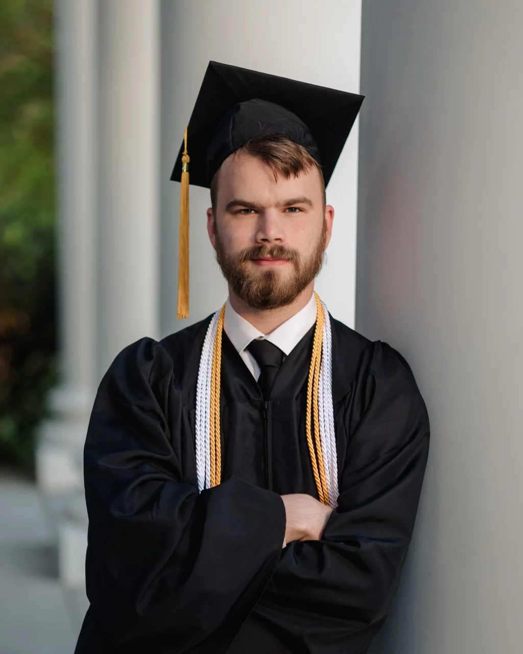 Patrick Henry College & Liberty University graduation photos.: A young man in a graduation cap and gown with honor cords stands with arms crossed, leaning against a column.