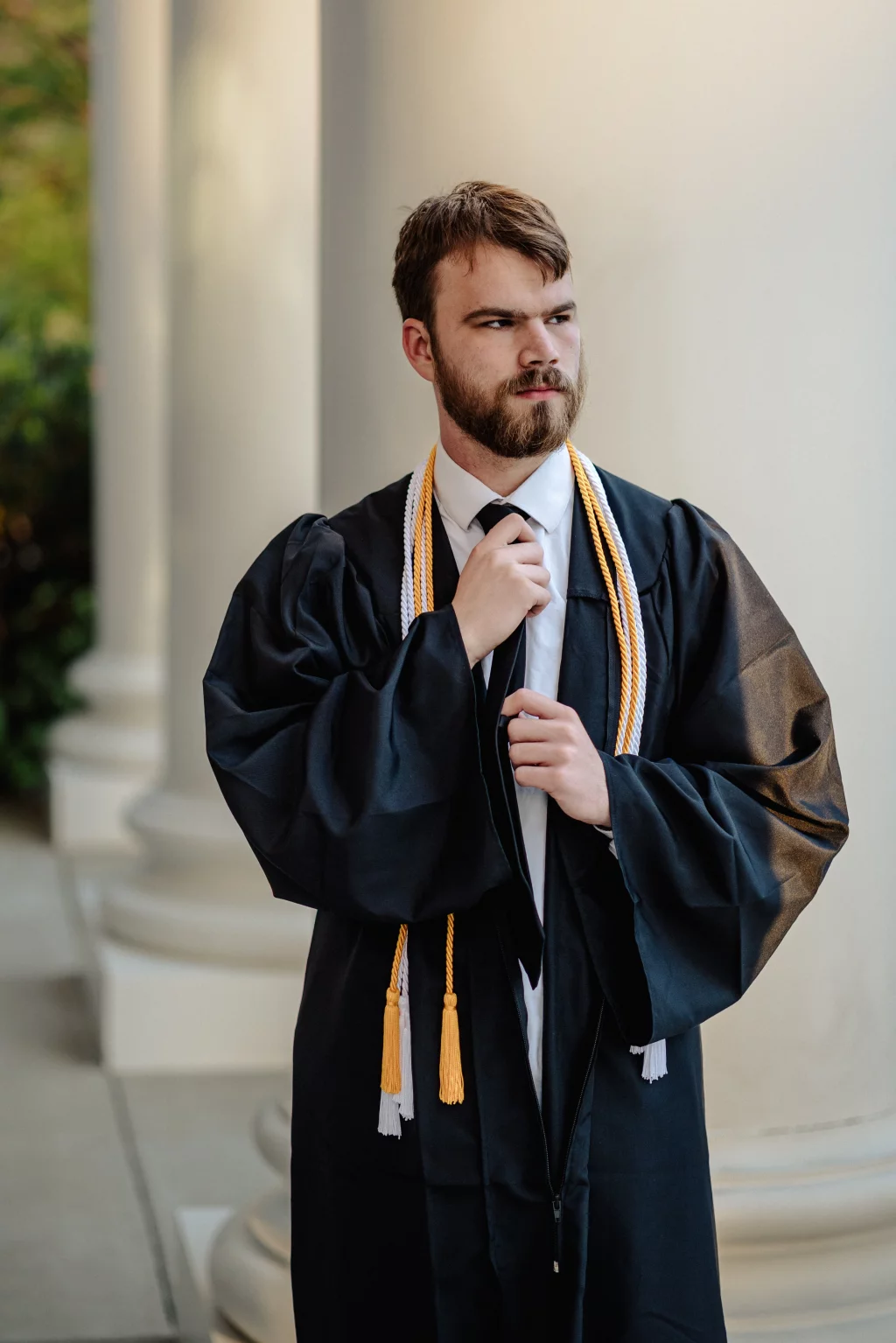 Patrick Henry College & Liberty University graduation photos.: A young man wearing a black graduation gown and honor cords adjusts his tie while standing in front of large white columns.