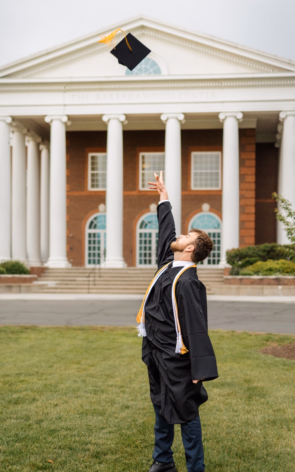 Patrick Henry College & Liberty University graduation photos.: A graduate in a black cap and gown tosses their graduation cap into the air in front of a large building with white columns.
