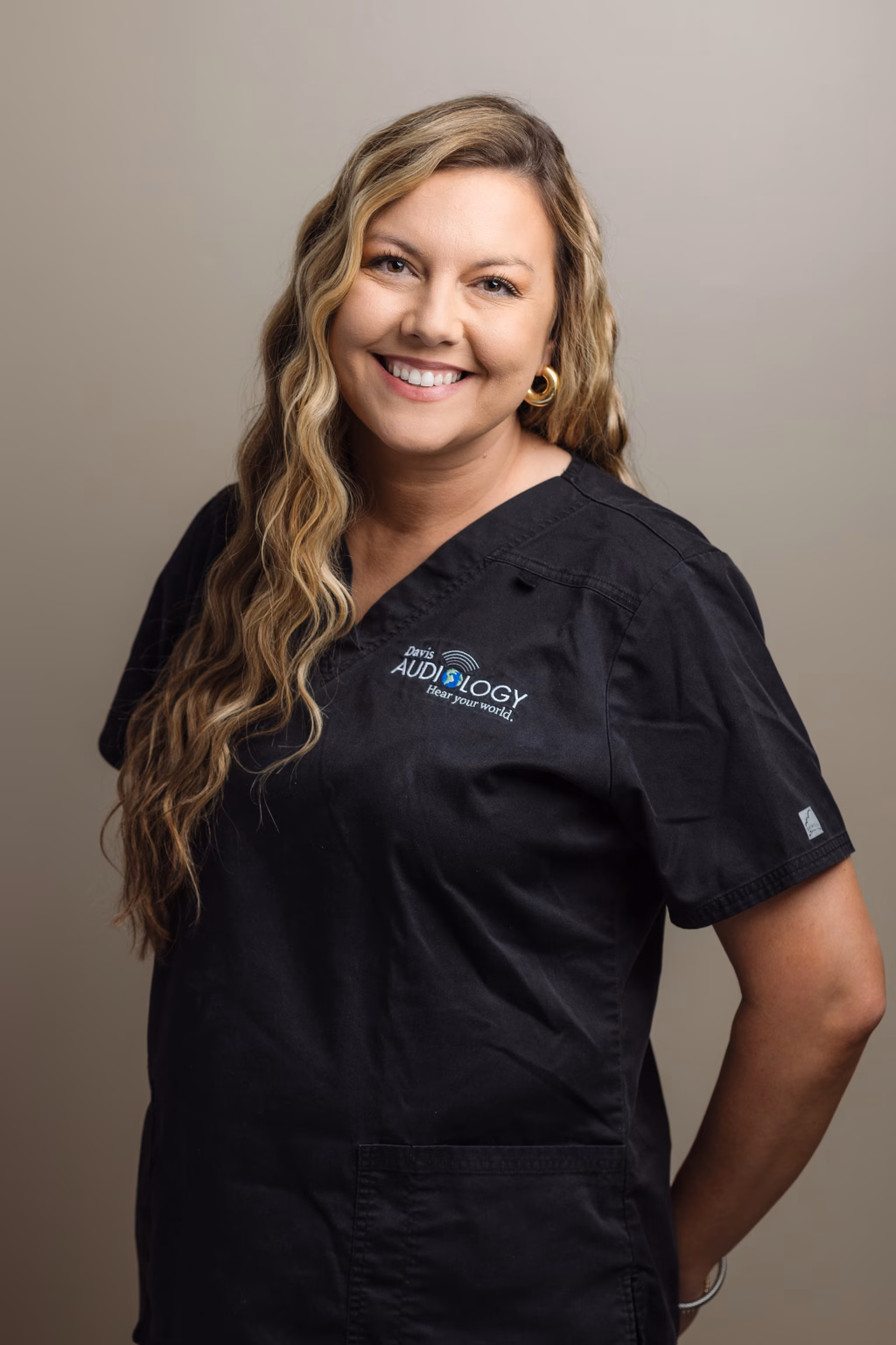 Headshots and branding for Davis Audiology in Taylors SC.: Smiling woman with long wavy blonde hair wearing black medical scrubs embroidered with &ldquo;Davis Audiology,&rdquo; posing against a neutral background.